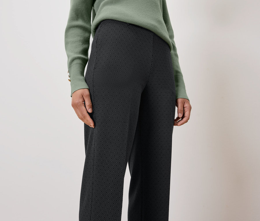 Žena má na sobě zelený svetr a černé vzorované kalhoty.