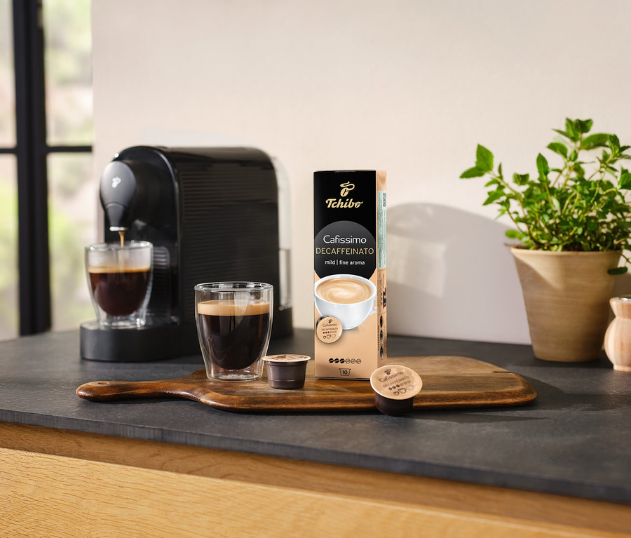 Kávovar nalévá kávu do sklenice vedle balení kapslí Cafissimo Decaffeinato.
