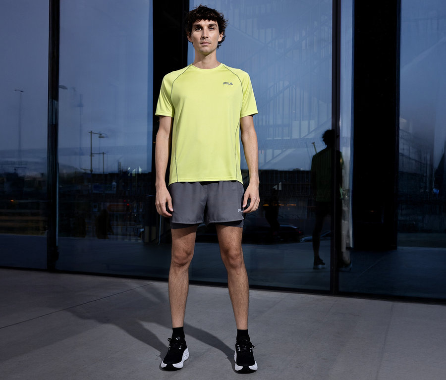 Muž stojí před budovou a má na sobě žluté tričko Fila, šedé šortky a černé boty.