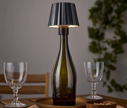 Stolní lampa ve tvaru lahve vína stojí na dřevěném stole vedle dvou sklenic na víno a misky ořechů.