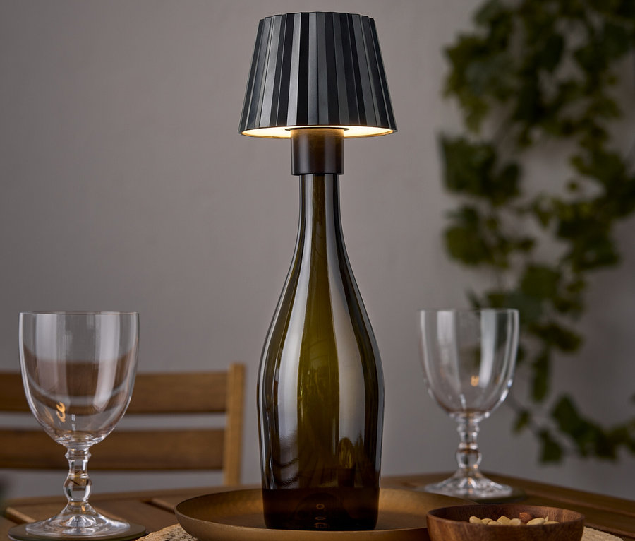 Stolní lampa ve tvaru lahve vína stojí na dřevěném stole vedle dvou sklenic na víno a misky ořechů.