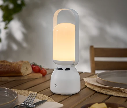 Zapnutá, přenosná, bílá lampa s rukojetí stojí na dřevěném stole vedle talíře, příboru, chleba, rajčat a sýra.