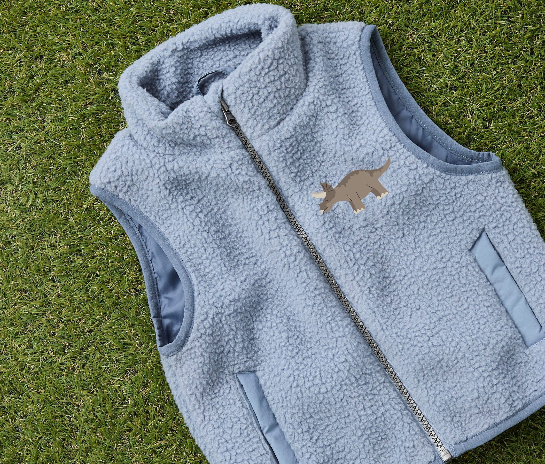 Světle modrá fleecová vesta s motivem triceratopse leží na zelené trávě.