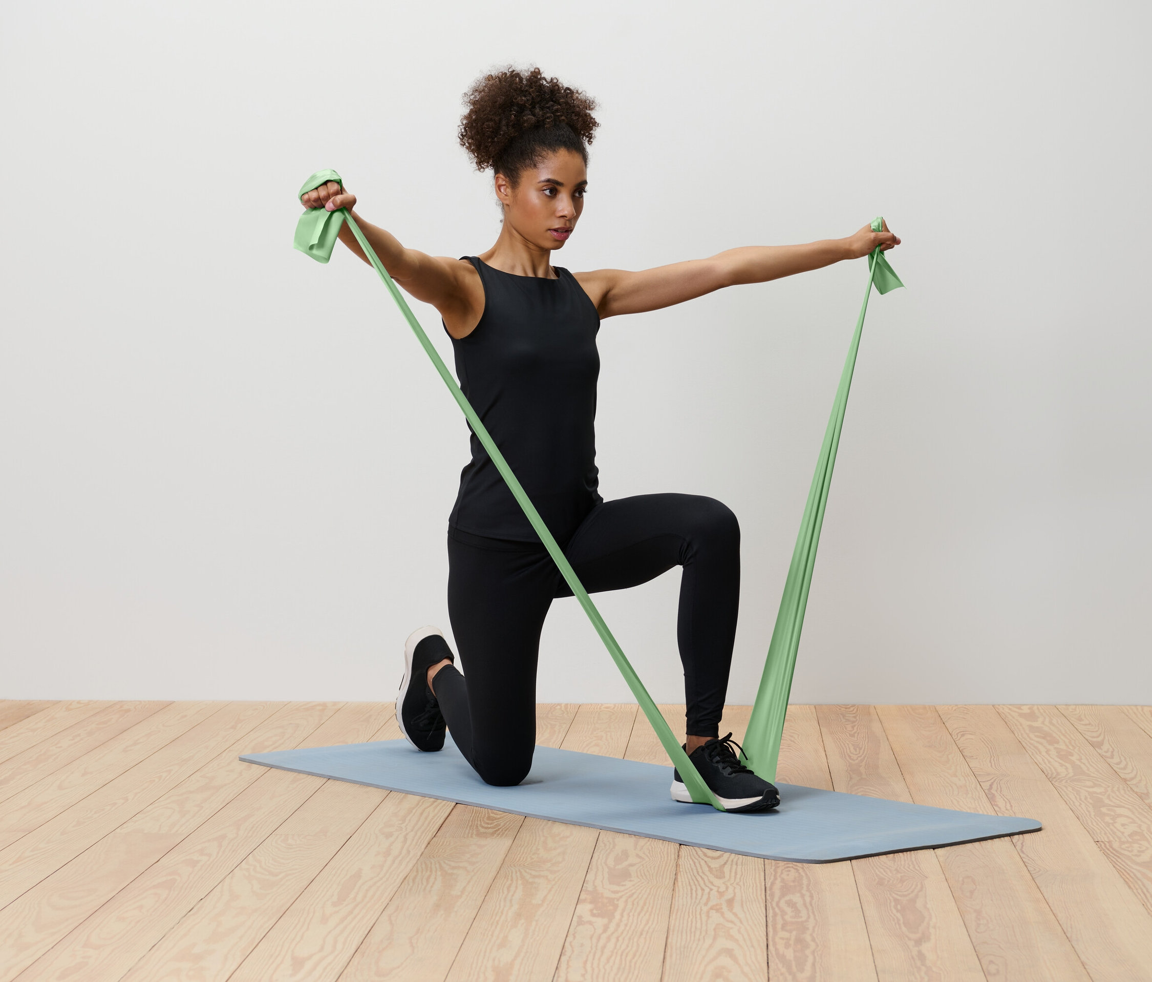 Žena klečí na modré podložce na jógu a natahuje zelenou odporovou gumu.