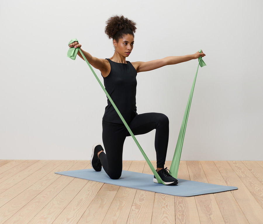 Žena klečí na modré podložce na jógu a natahuje zelenou odporovou gumu.