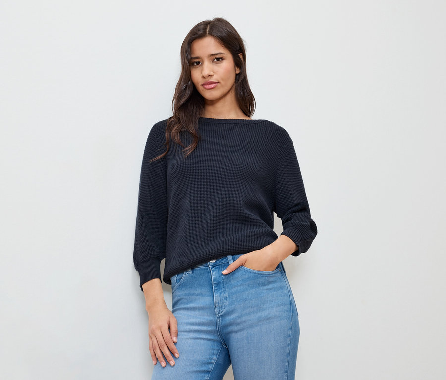 Žena s rukou v kapse má na sobě tmavě modrý jemný pletený svetr.