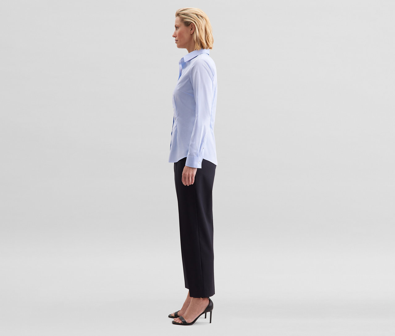 Žena stojí z profilu, má na sobě světle modrou halenku slim fit non iron Seidensticker, černé kalhoty a černé sandály na podpatku.