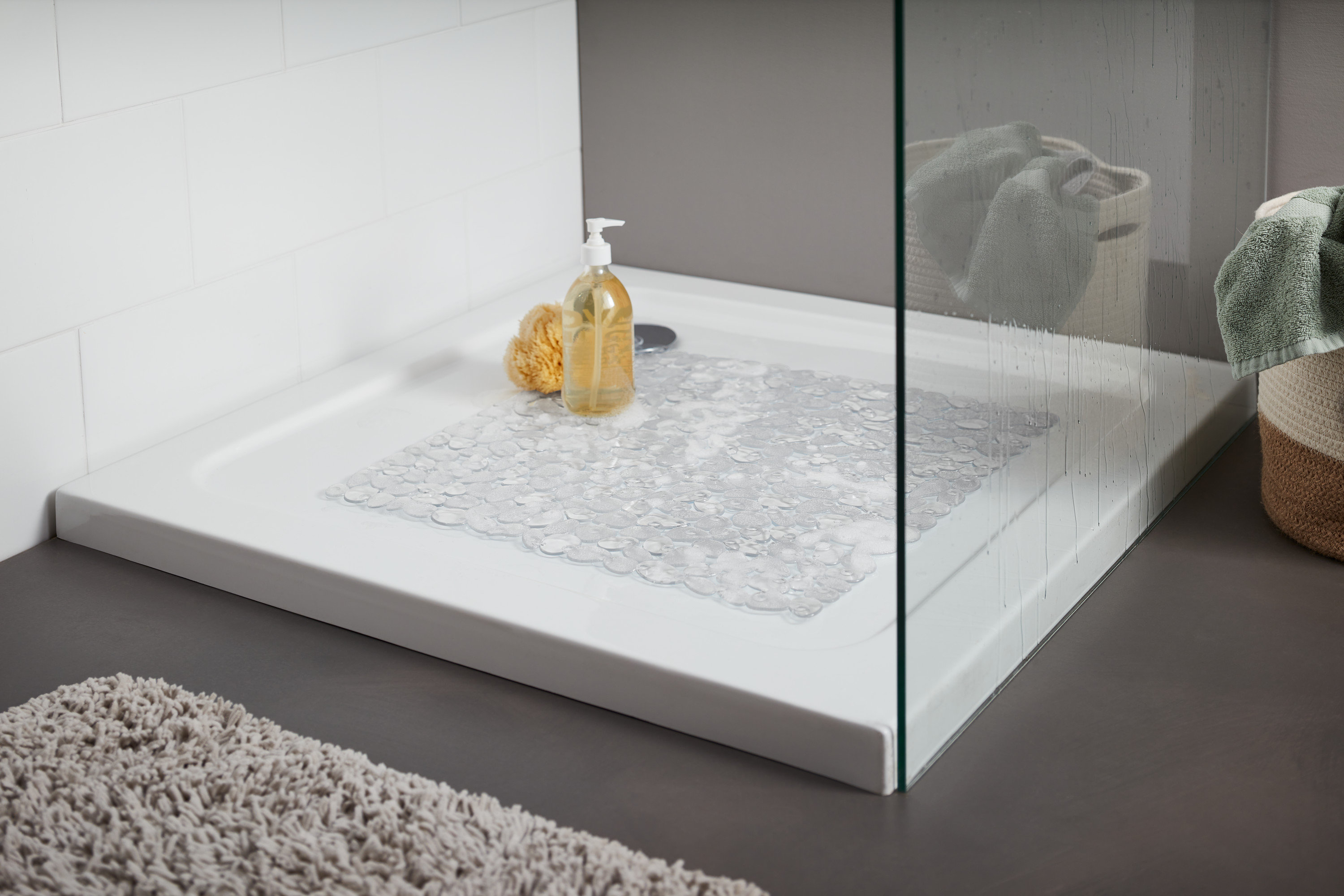 Vanička se sprchovým koutem, v ní je položena průhledná podložka do sprchového koutu. Na vaničce je žlutá mycí houba a lahvička s pumpičkou.