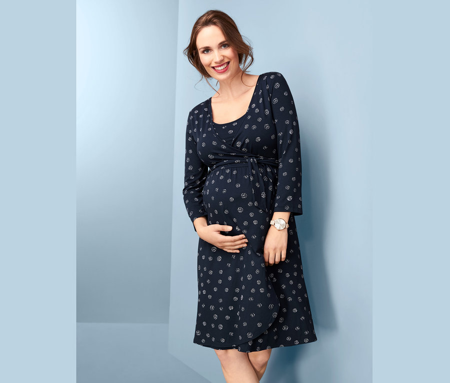 Usmívající se těhotná žena drží břicho a má na sobě tmavě modré zavinovací těhotenské šaty s potiskem.