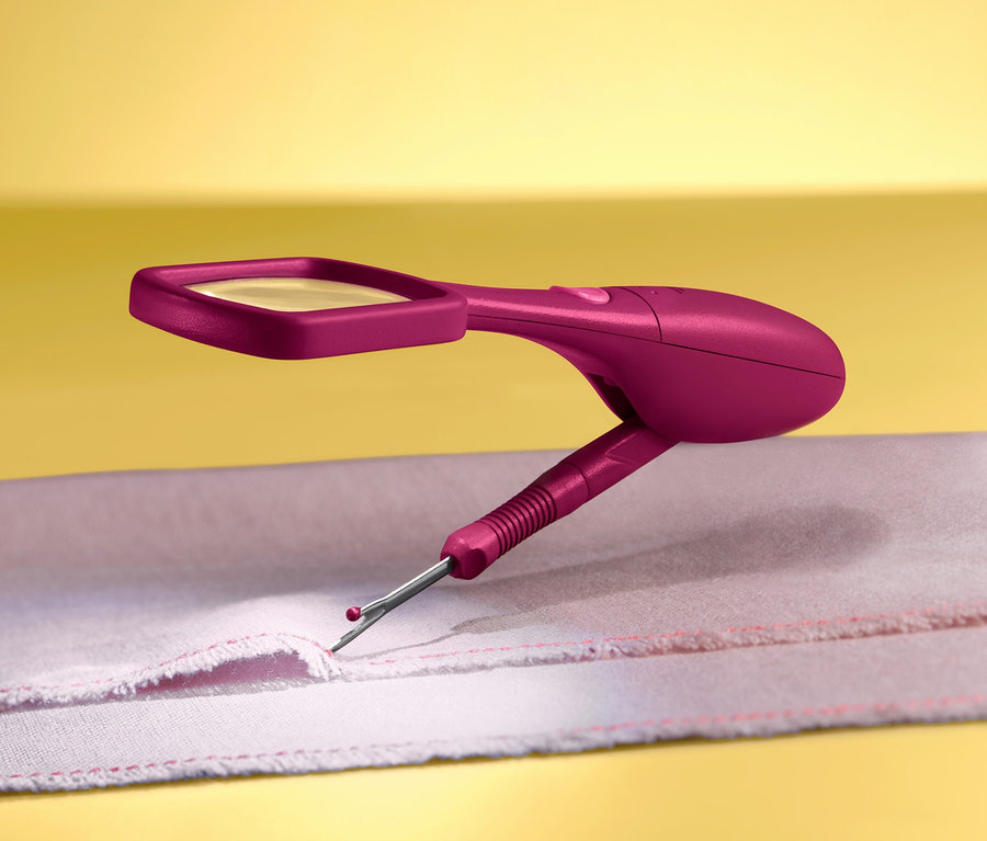 Detailní záběr růžového páráčku s lupou a LED osvětlením, který se používá na látce s červenou nití na žlutém pozadí.