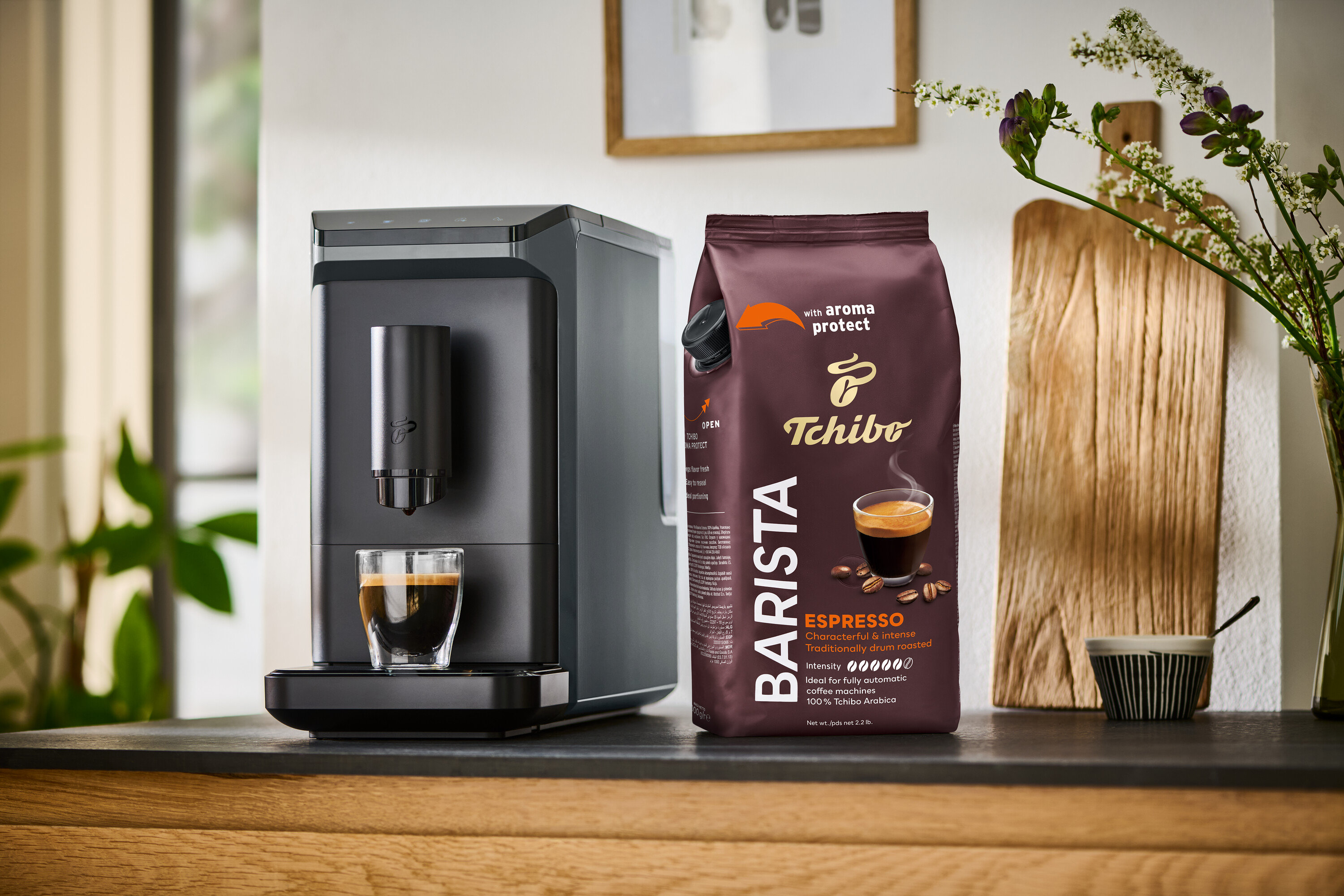 Automat na kávu připravuje kávu do sklenice vedle balení zrnkové kávy Barista Caffè Crema.