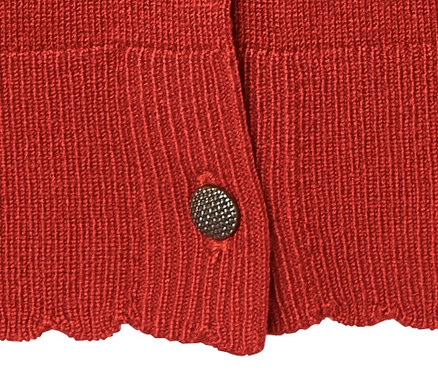 Detail červeného kabátku z jemné pleteniny s ozdobným knoflíkem.
