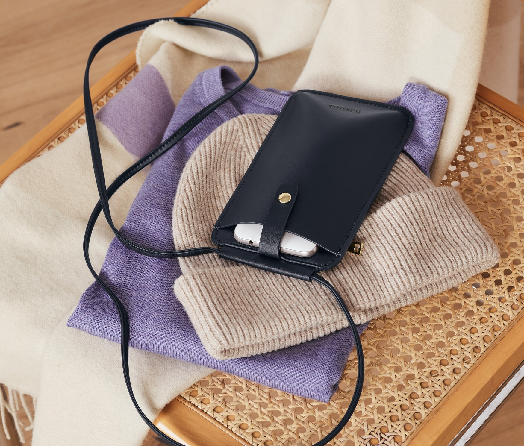 Na proutěné židli leží tmavě modrá taštička na mobil, béžová čepice, fialový svetr a pruhovaná šála.