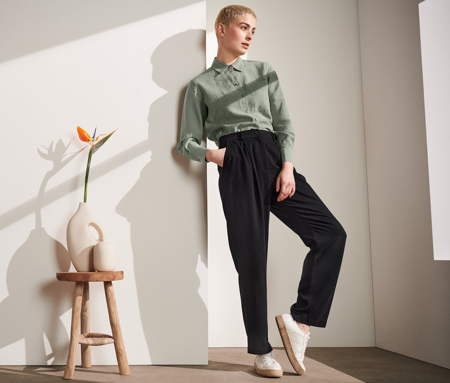 Žena opřená o zeď v zelené košili a černých kalhotách s puky.