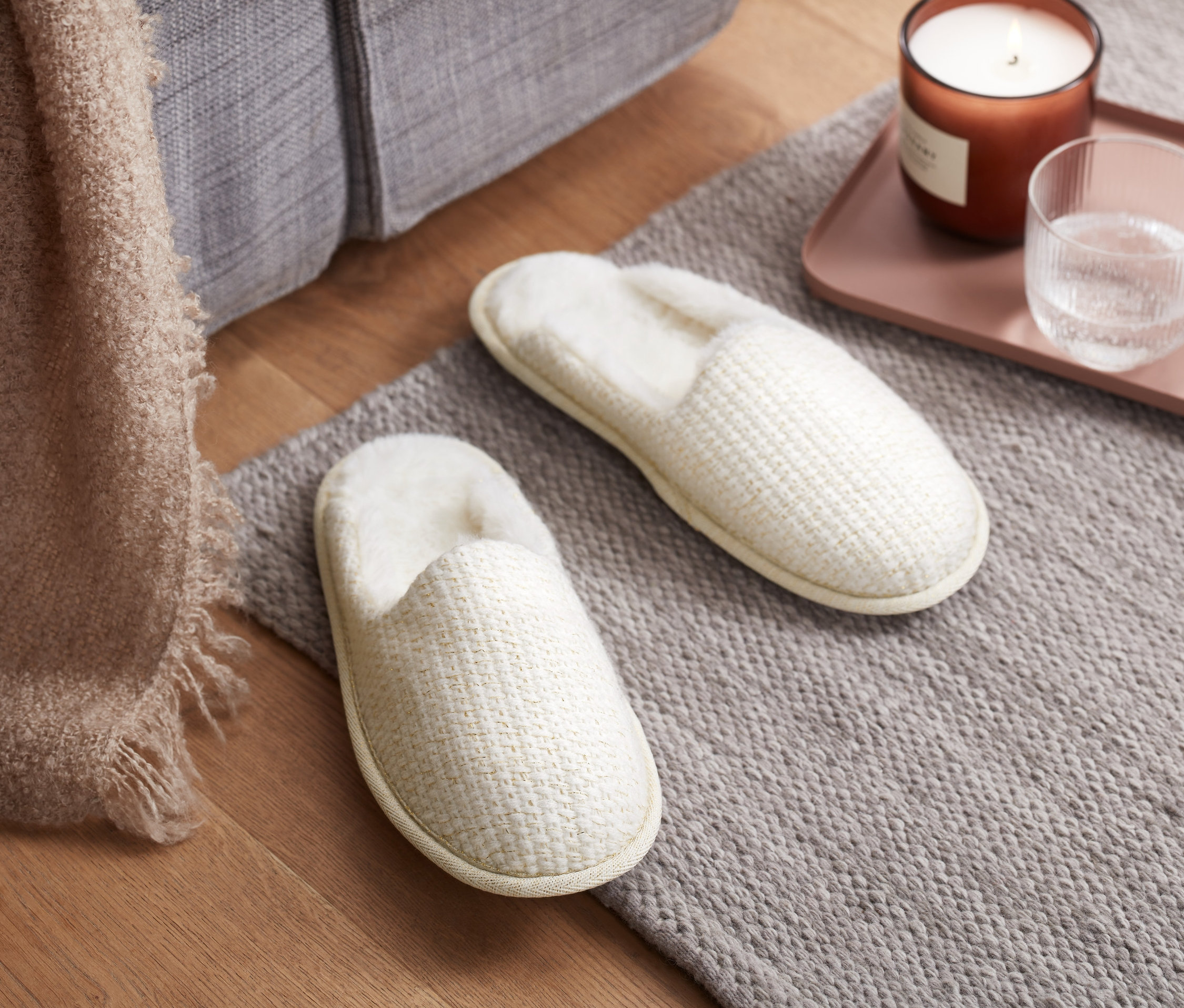 Dvoje pantofle leží na šedém koberci vedle podnosu se svíčkou a sklenicí vody.
