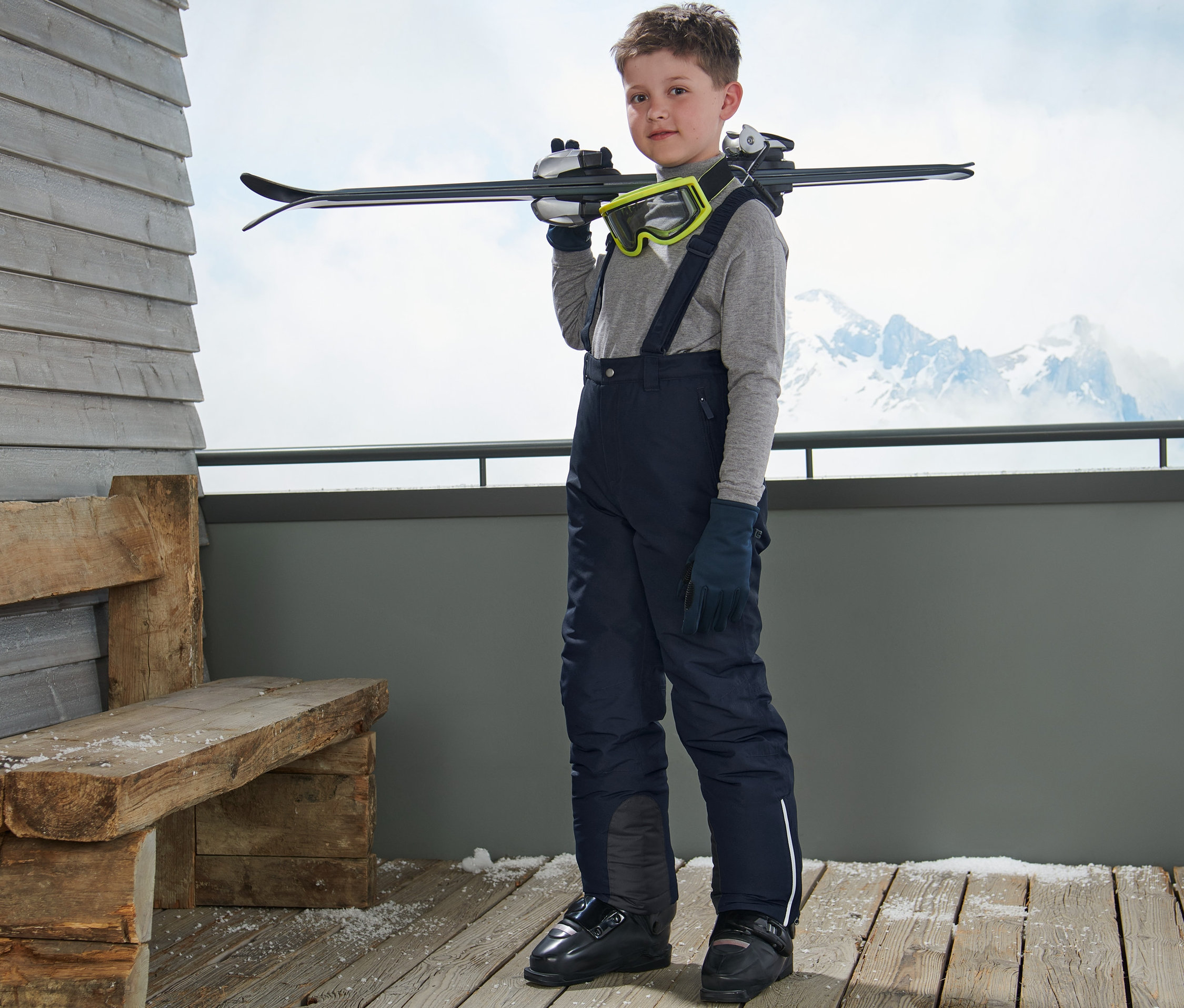 Chlapec stojí a přes rameno si nese lyže. Má na sobě lyžařské kalhoty, tmavě modré, šle, lyžařské brýle a rukavice.