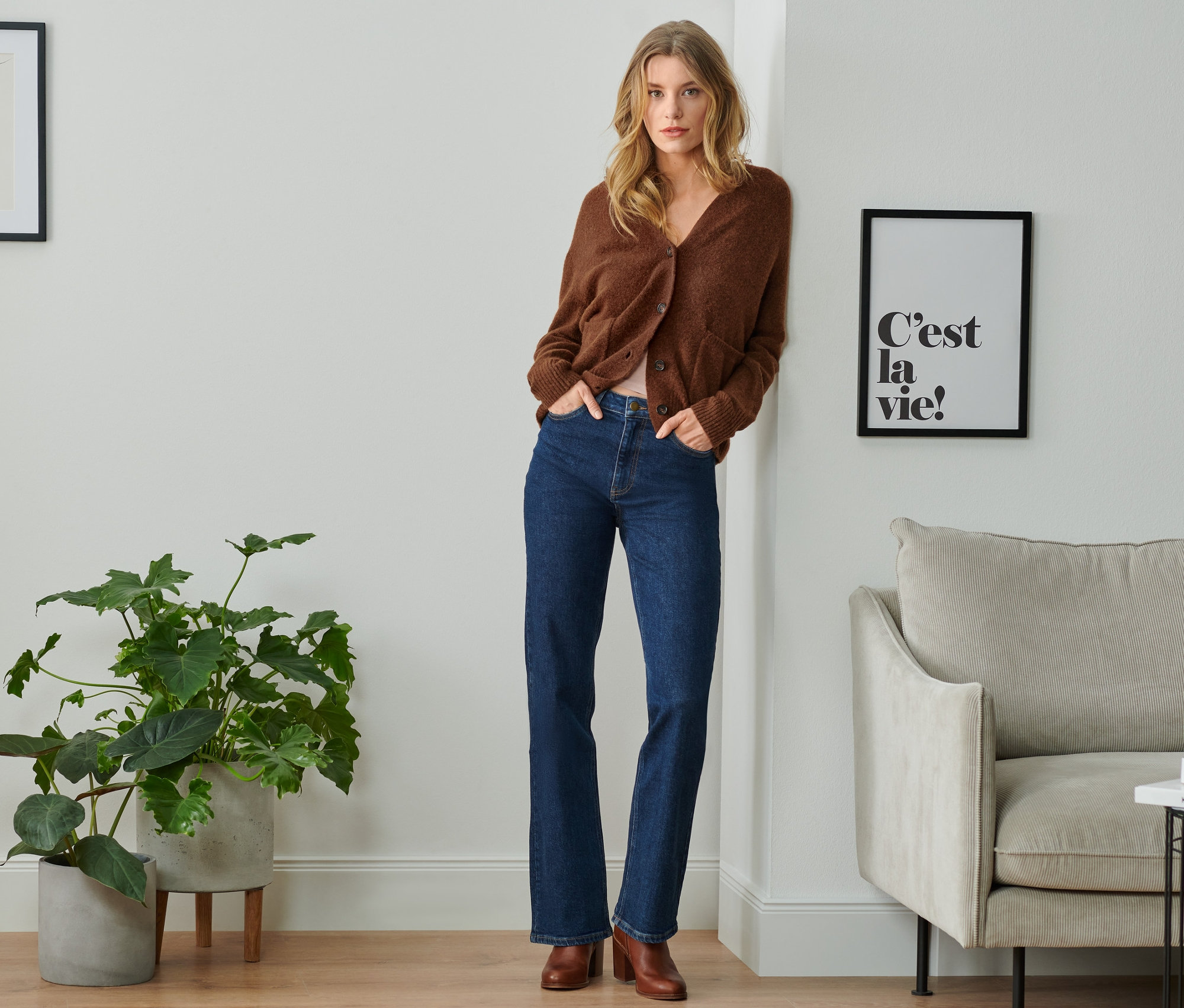 Žena opřená o zeď má na sobě hnědý kardigan a džíny bootcut.