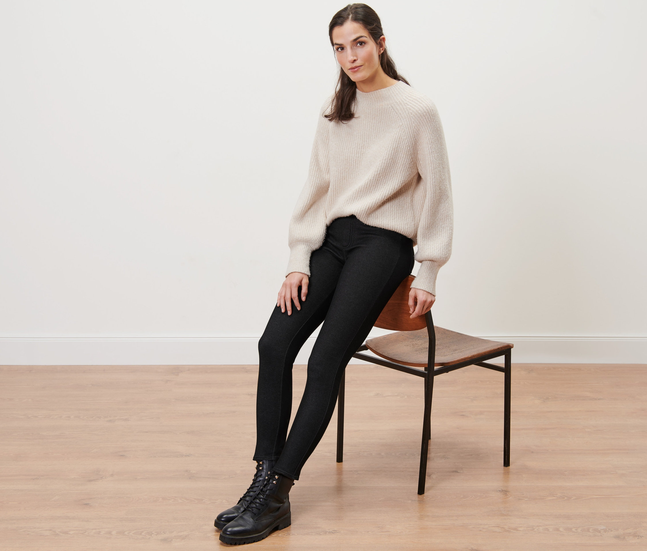 Žena sedí na židli a má na sobě legíny v džínovém vzhledu s podšívkou, svetr a boty.