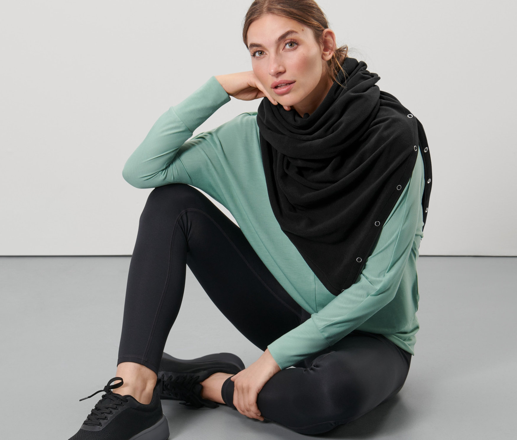 Žena sedí na podlaze a má na sobě černou oversize šálu 3 v 1.