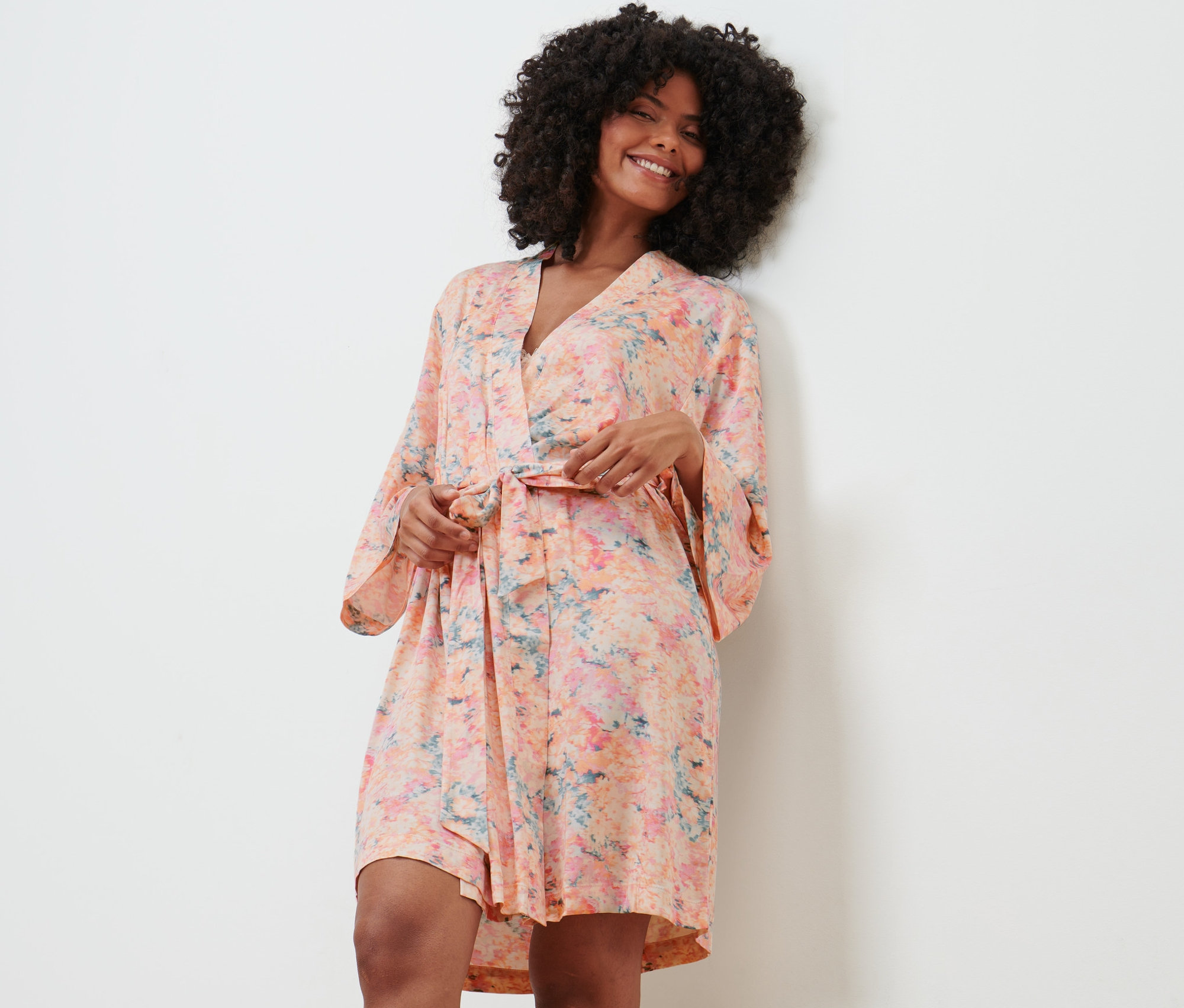 Žena v županu stojí opřená o zeď a usmívá se.