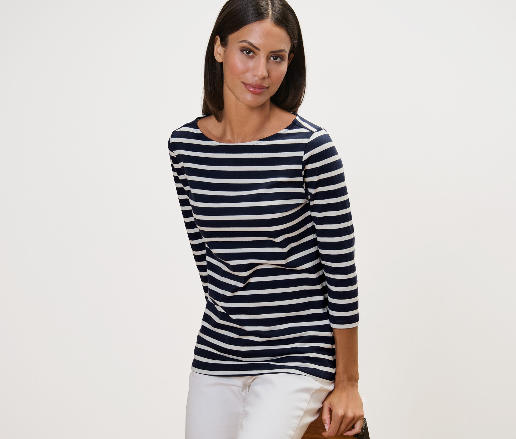 Žena pózuje v proužkovaném triku s tříčtvrtečním rukávem v námořnické modré barvě a bílých kalhotách.