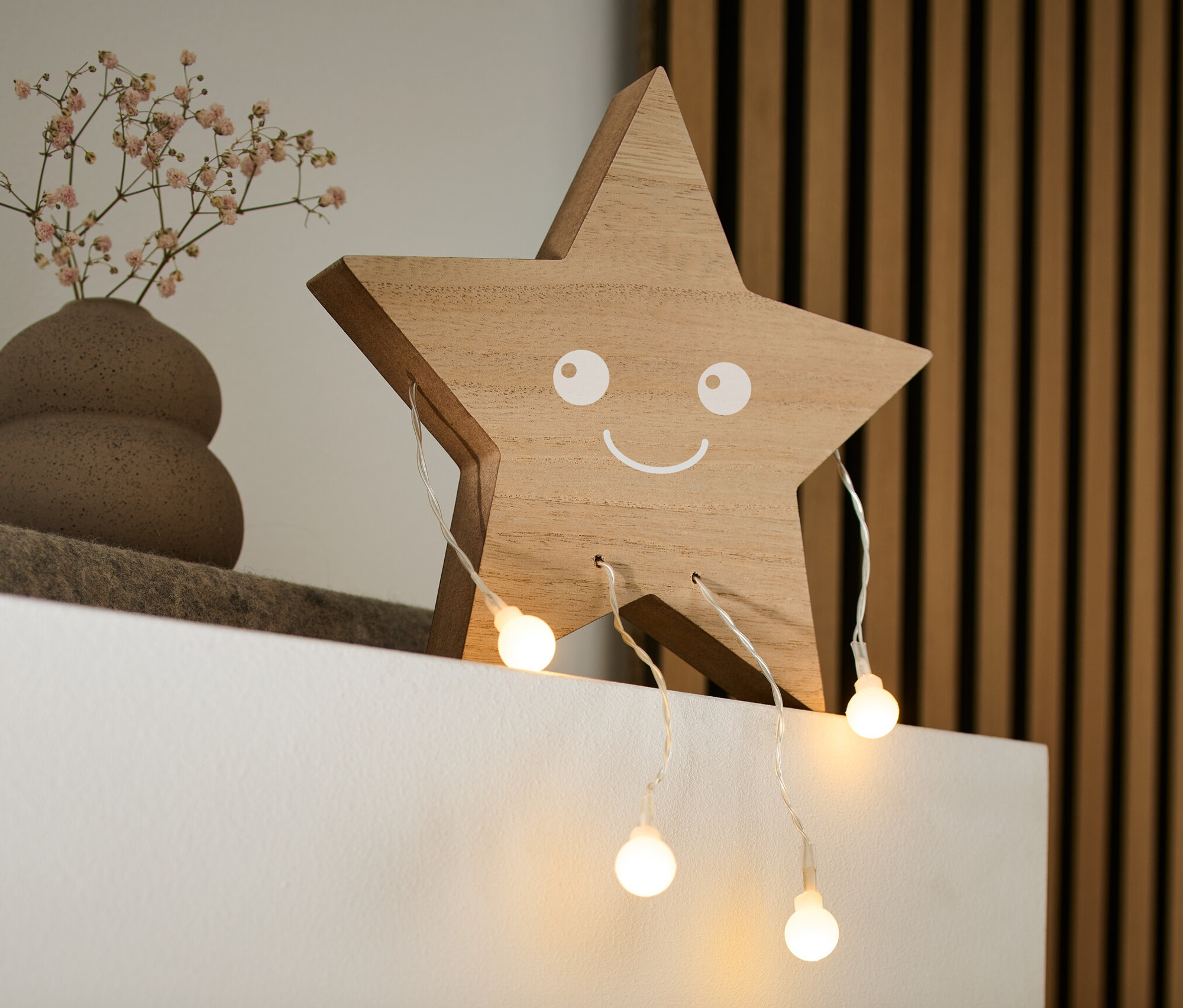 Na bílé polici stojí dřevěná LED dekorace Hvězda s usměvavým obličejem, váza s malými květy a kniha.