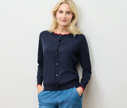 Žena má na sobě svetr z jemného úpletu se zvlněným okrajem a modré kalhoty a stojí u zdi.