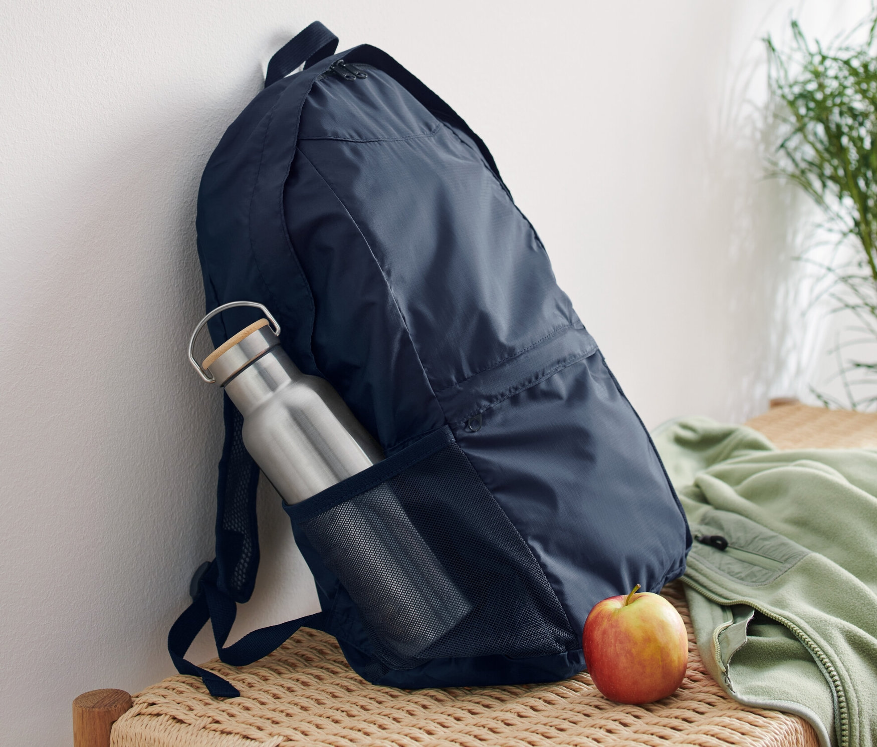 Tmavě modrý skládací batoh opřený o bílou zeď na proutěné lavici. V boční kapse batohu je stříbrná láhev na vodu a vedle batohu leží jablko.