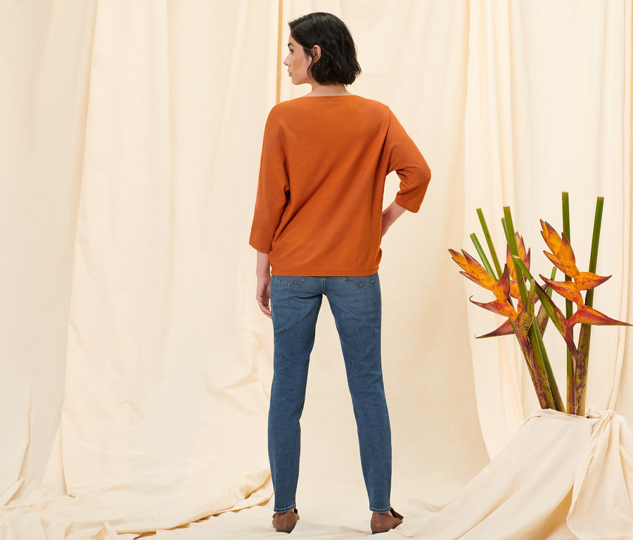 Žena stojí zády k fotoaparátu a má na sobě džíny slim »Fit Emma«, středně modrý denim, oranžový svetr z jemné pleteniny a boty z velurové kůže.