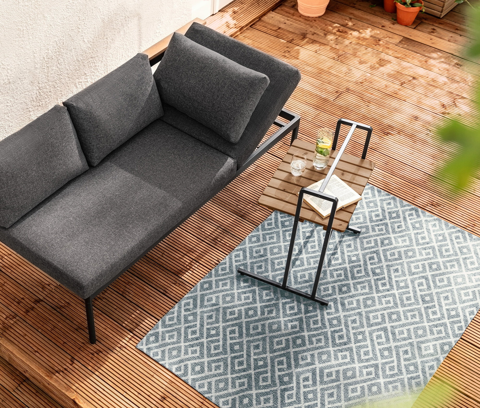 Pohled shora na jídelní lavici a lehátko Elin 2 v 1 vedle stolku ve tvaru C s velkou deskou na dřevěné terase.