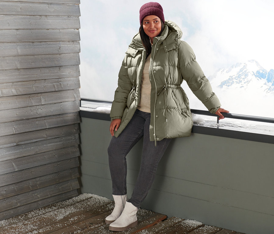 Žena se opírá o zábradlí v péřové bundě, džínách slim fit, kotníkových botách z velurové kůže a pletené čepici vínově červené.