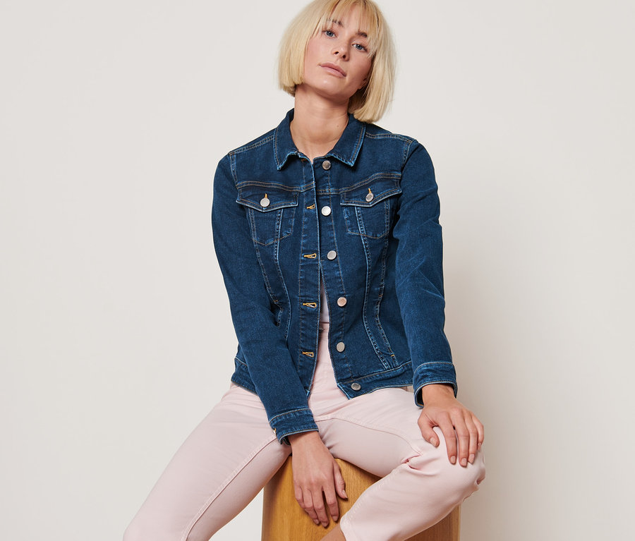 Žena sedí na dřevěné stoličce a má na sobě džínovou bundu středně modré barvy.