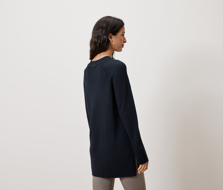 Žena stojí bokem a má na sobě tmavě modrý svetr a kostkované kalhoty.