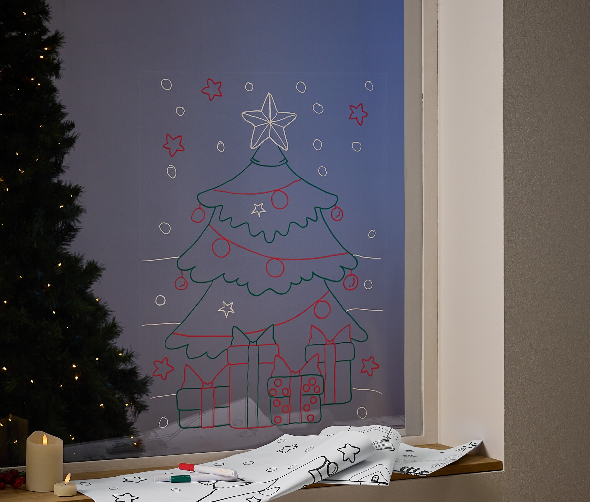 Na okně je nakreslený vánoční stromeček s ozdobami a dárky. Na parapetu leží papír s vánočními motivy a fixy.