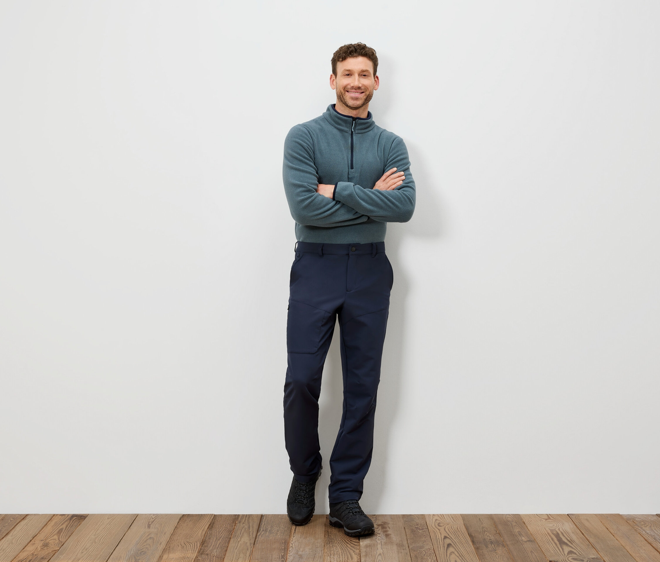 Muž stojí s rukama zkříženýma na hrudi, má na sobě modrozelený pulovr a tmavě modré kalhoty.