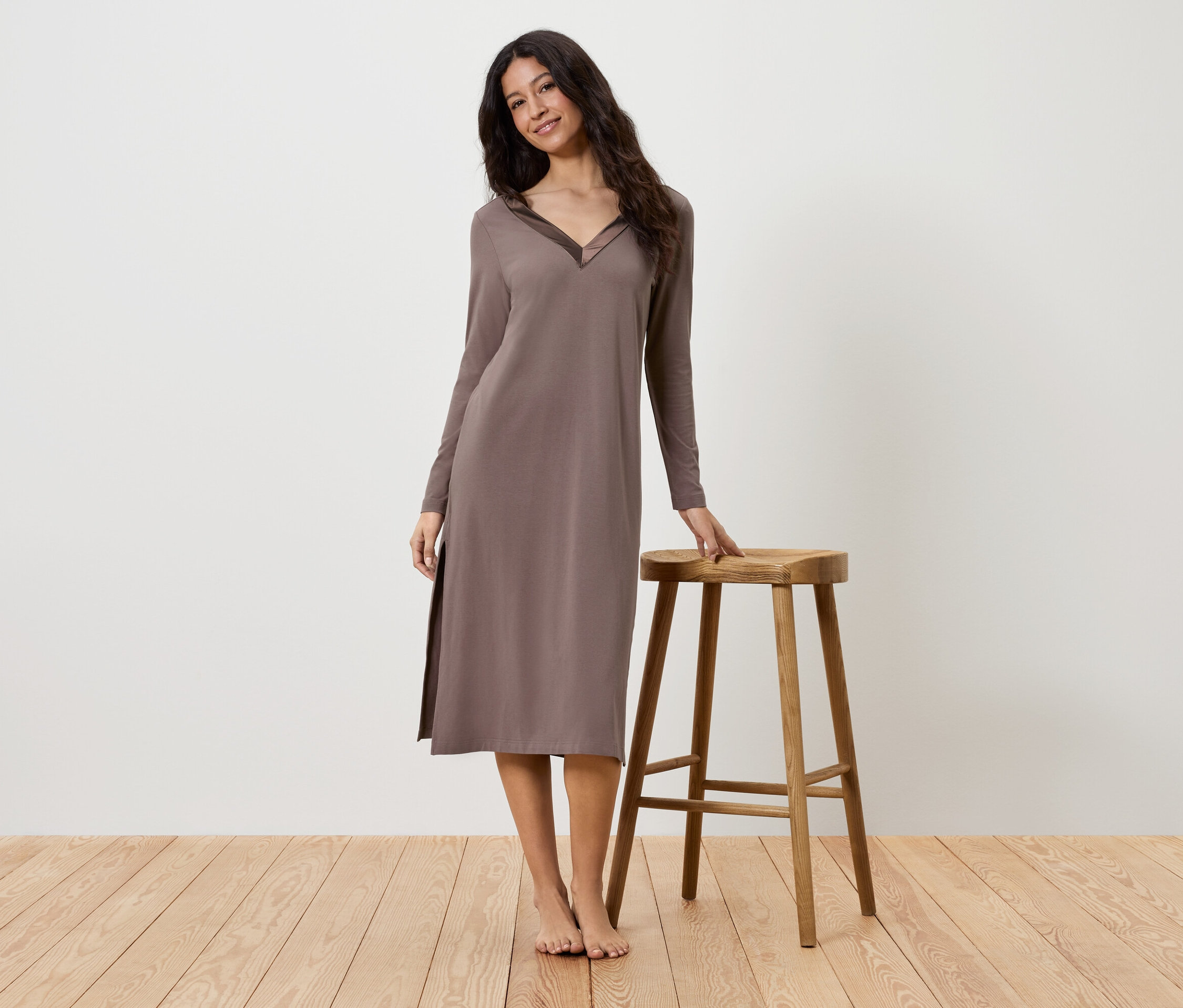 Žena v hnědých šatech s dlouhým rukávem stojí vedle dřevěné stoličky.