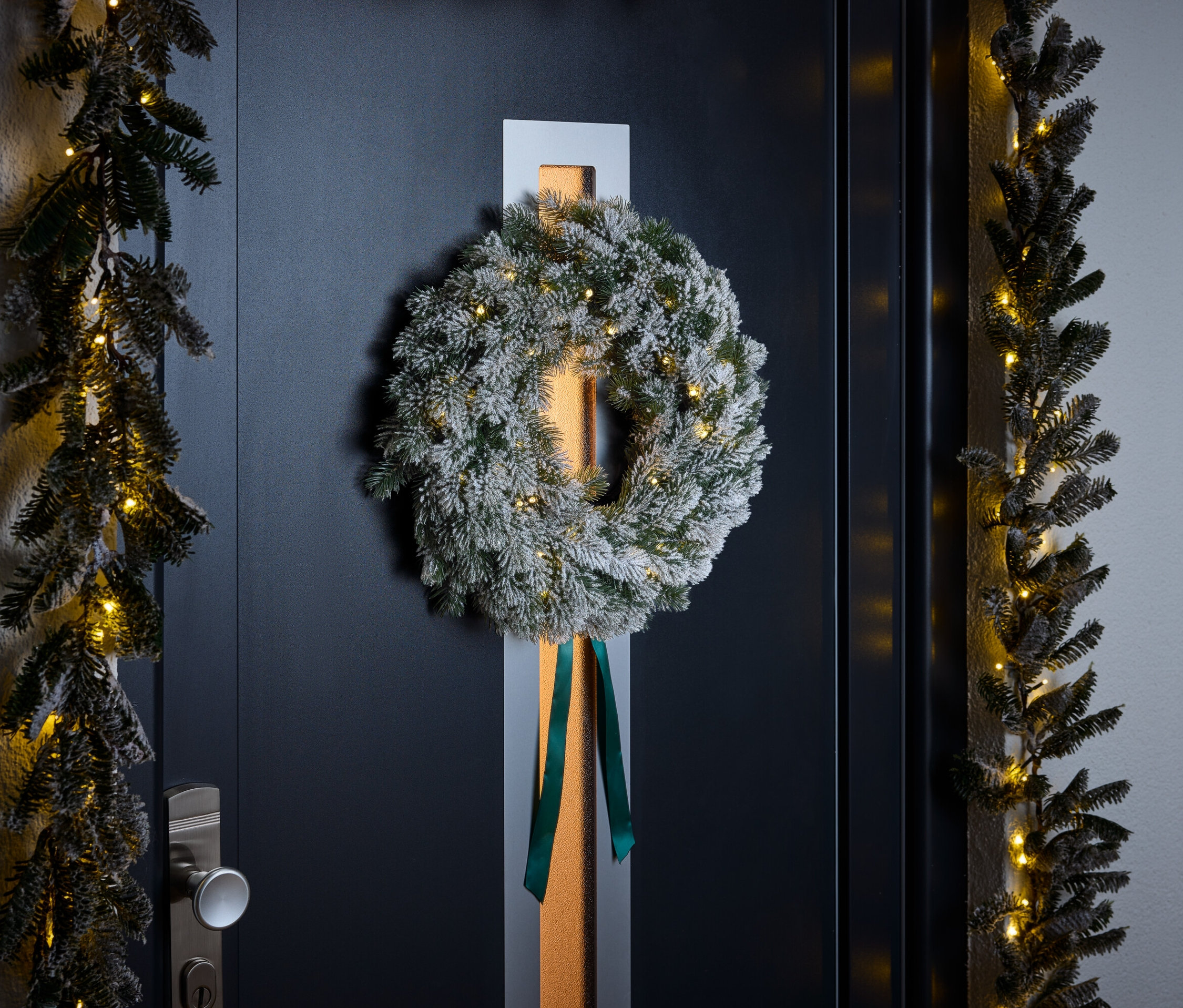 Věnec s bílým posypem a zelenou stuhou visí na tmavě modrých dveřích. Girlandy s malými světýlky lemují dveře.