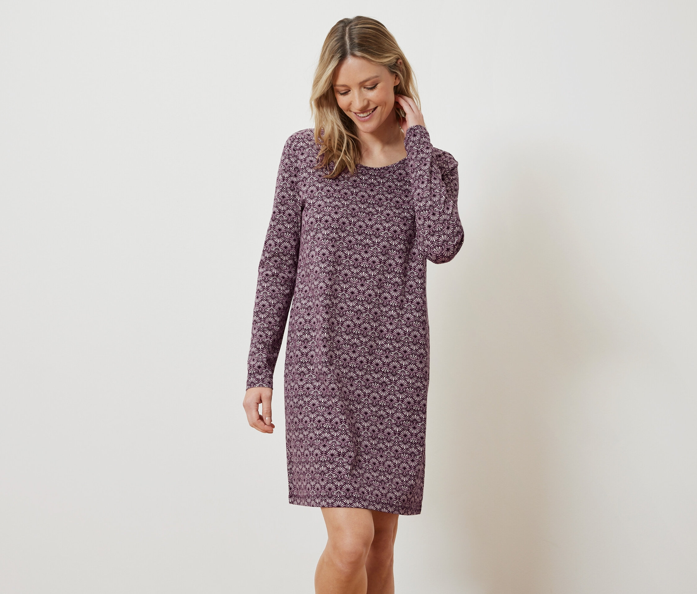 Žena s blond vlasy a úsměvem pózuje v dlouhých šatech s květinovým vzorem.