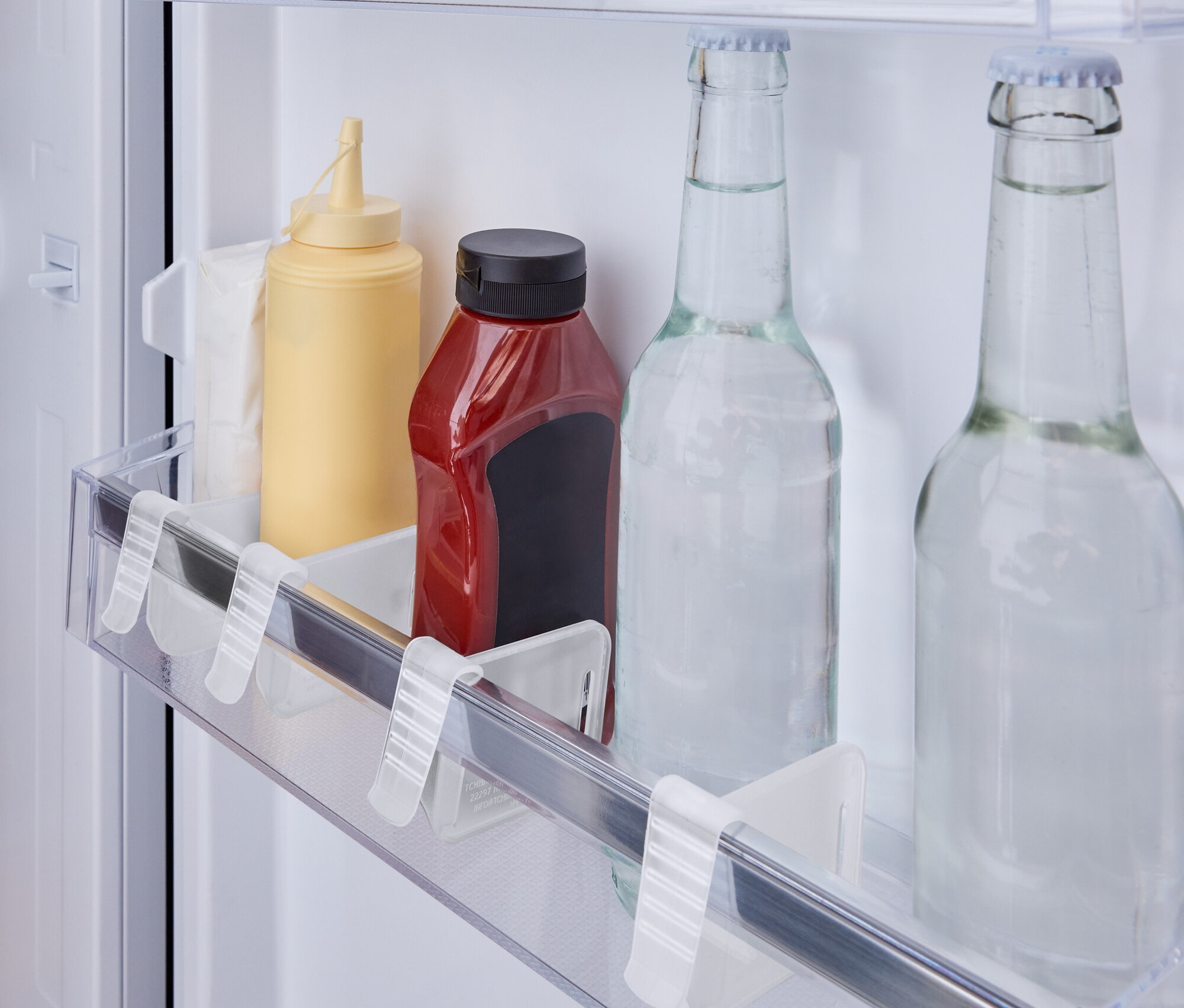 V lednici na poličce stojí žlutá láhev s hořčicí, červená láhev s kečupem a dvě skleněné lahve s vodou.