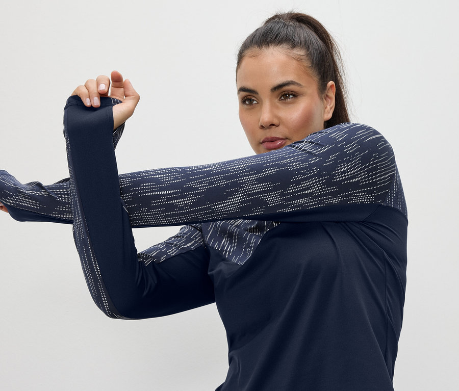 Žena protahuje paže v tmavě modrém sportovním topu s dlouhým rukávem a otvory pro palce.