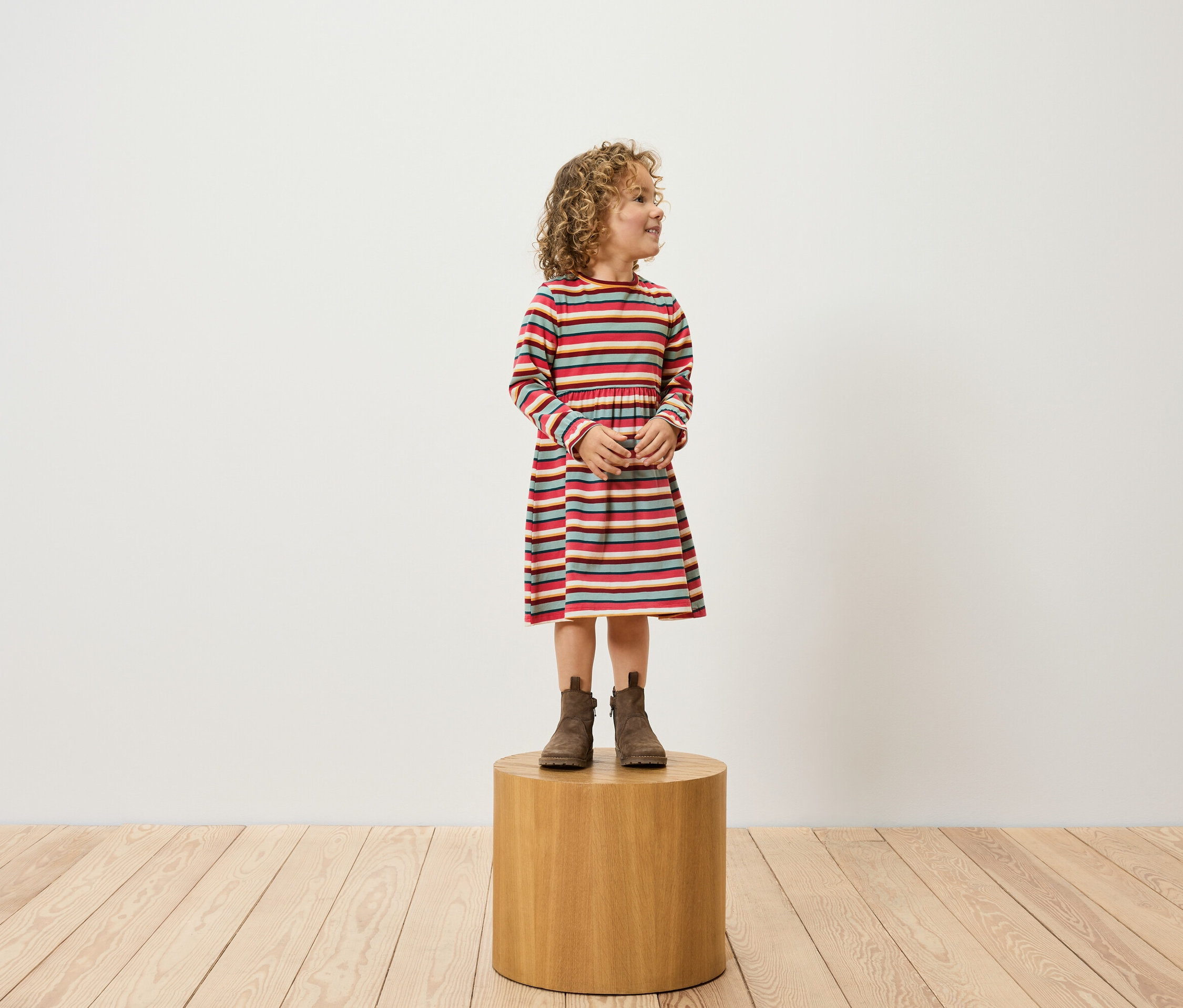 Dívka s kudrnatými vlasy stojí na dřevěném podstavci a má na sobě pruhované šaty a hnědé boty.