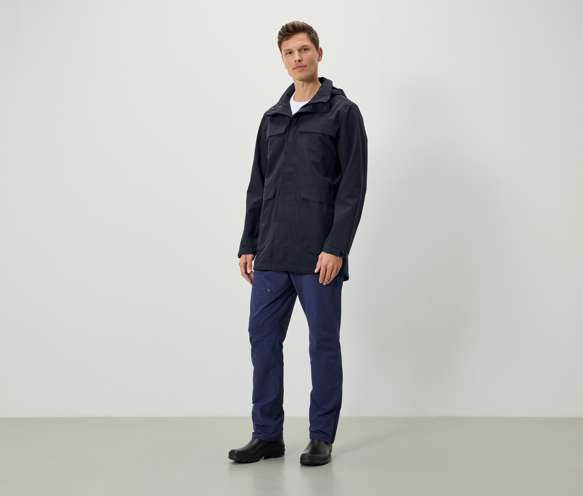 Muž stojí a má na sobě tmavě modrou bundu s kapucí, tmavě modré kalhoty a černé boty.