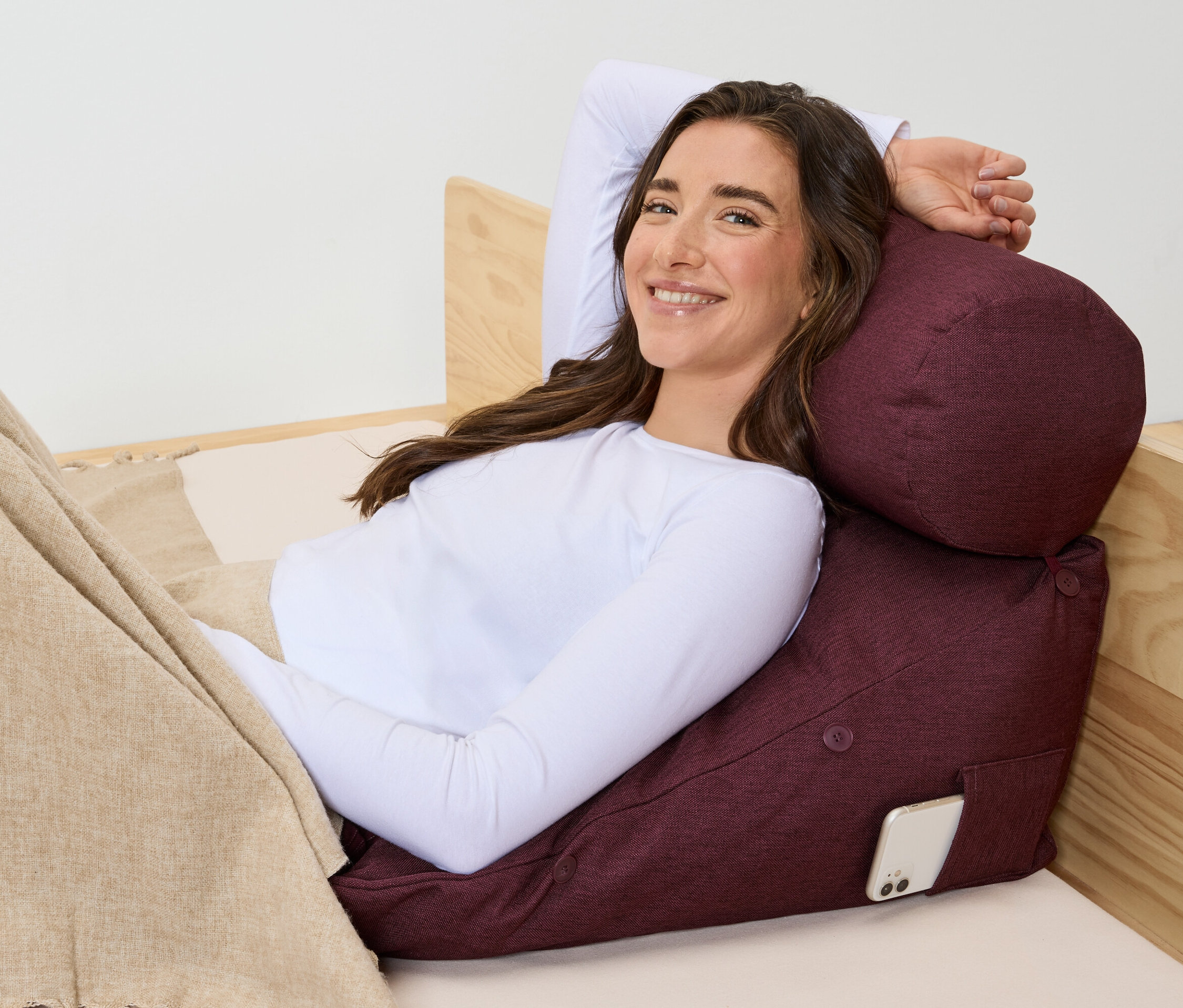 Žena leží na posteli a opírá se o bordó polštář s telefonem v kapse.