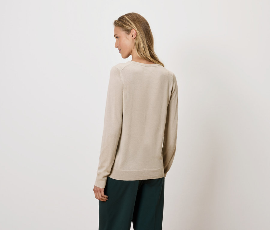 Žena se dívá přes rameno a má na sobě béžový svetr a zelené kalhoty.