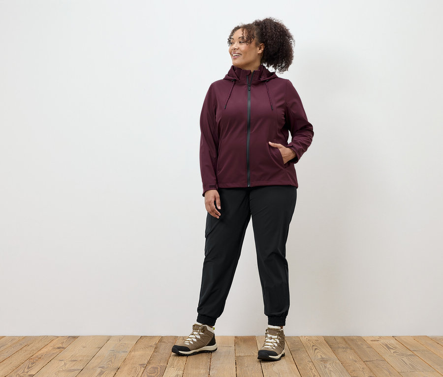 Žena v bordó bundě na zip, černých kalhotách a hnědých botách stojí na dřevěné podlaze.