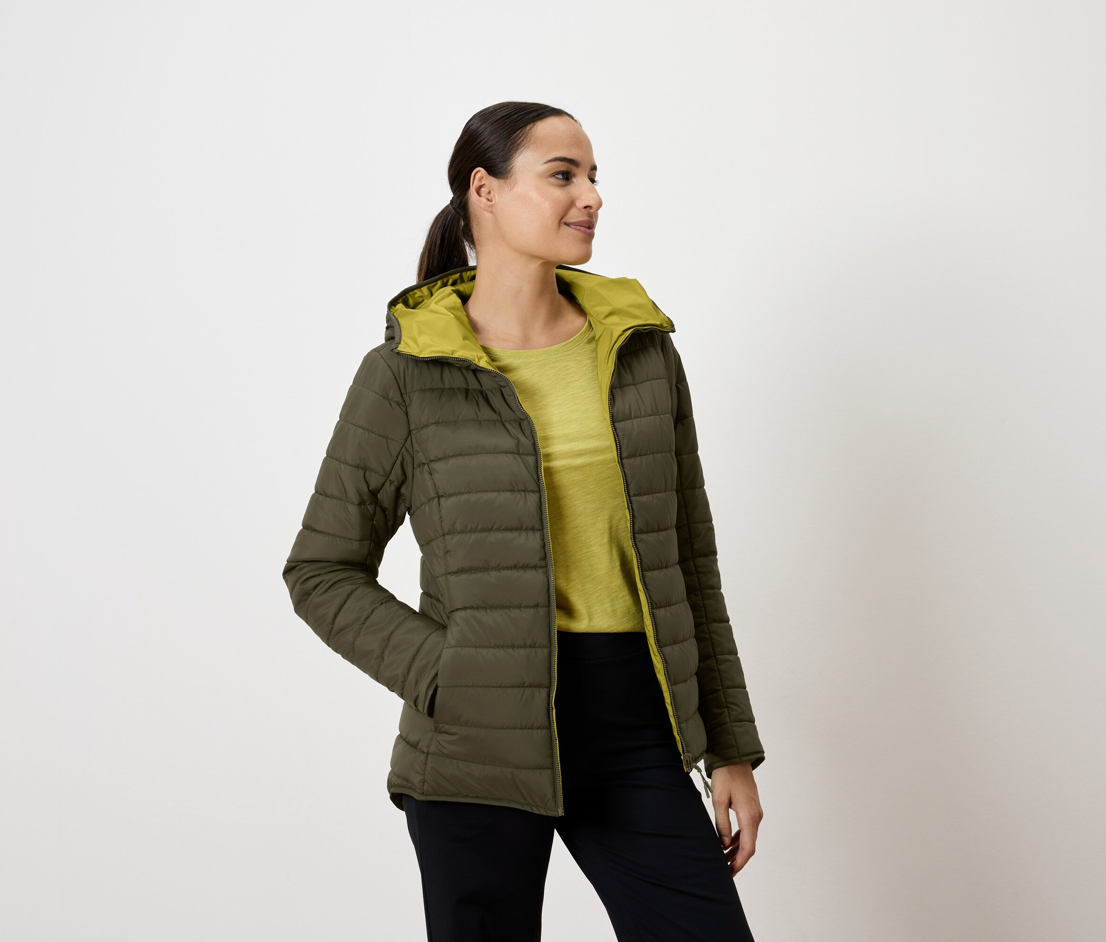 Žena v olivově zelené prošívané bundě s kapucí, ruce v kapsách, dívá se doprava.