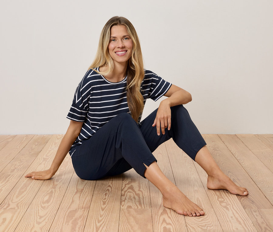 Blonďatá žena sedí na dřevěné podlaze v námořnickém pruhovaném tričku a tmavě modrých kalhotách.