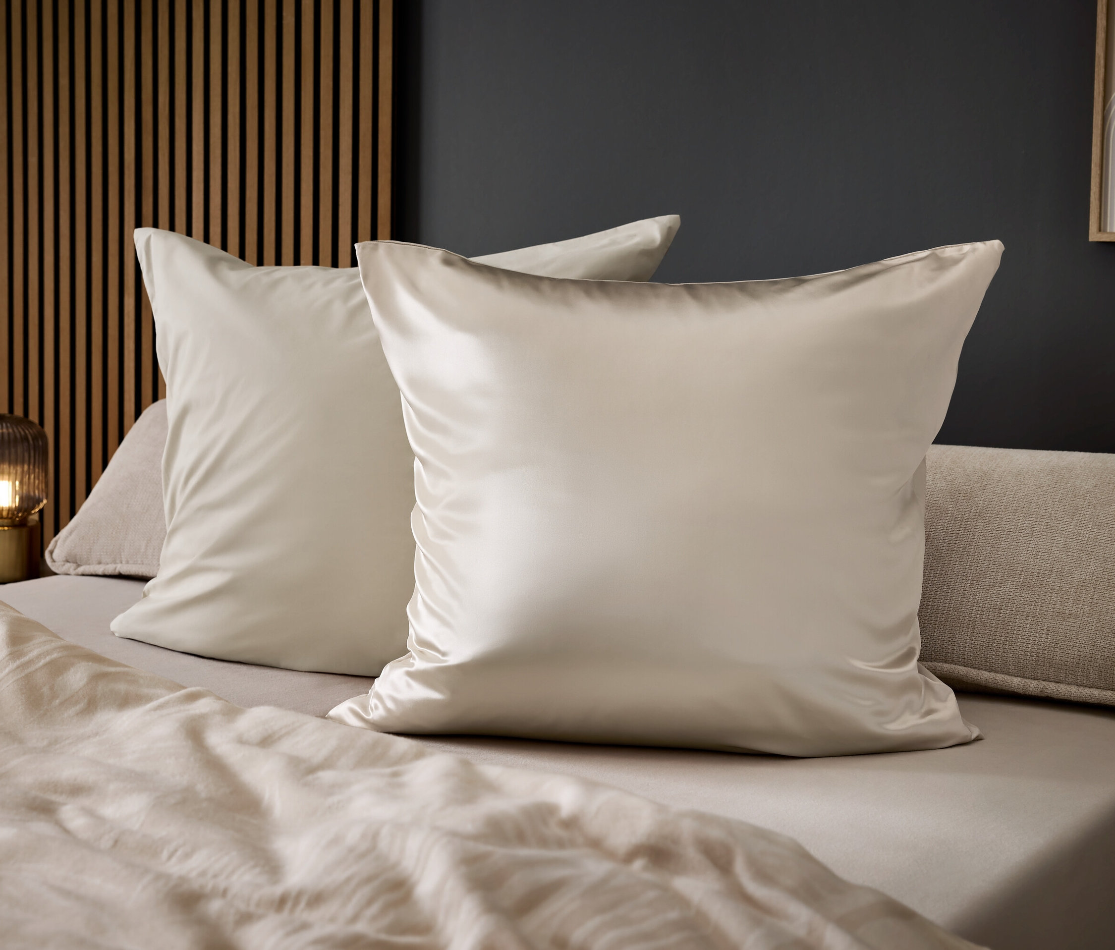 Dva lesklé béžové polštáře na posteli s béžovými prostěradly.