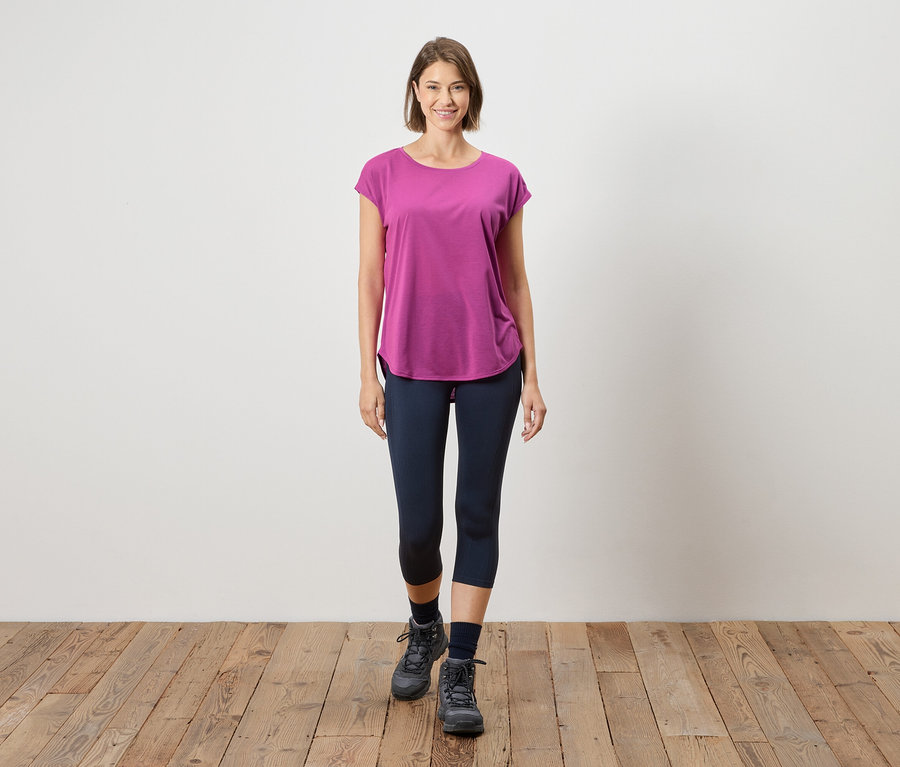 Žena stojí na dřevěné podlaze před bílou stěnou. Má na sobě růžové tričko, modré legíny a šedé boty.