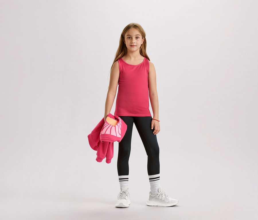 Dívka v růžovém tílku, černých legínách, bílých ponožkách a teniskách drží růžovou bundu a tašku.
